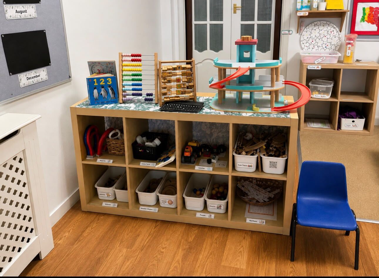 Open shelving with labelled tubs of magnets, loose parts, vehicles, animals, wooden shapes and numbers, a wooden marble run on top